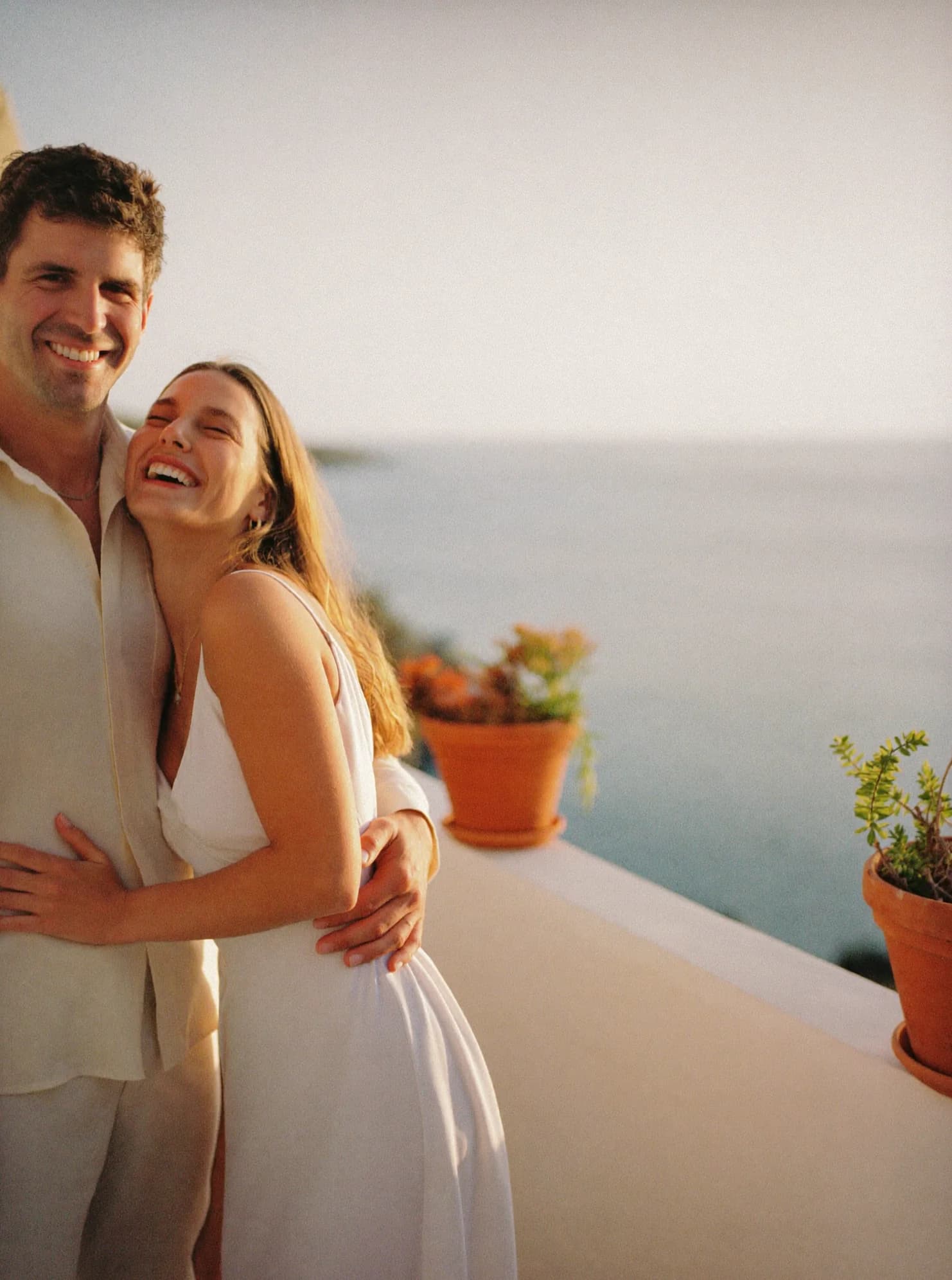 A couple in their forties laughing together on a sunlit seaside terrace, his arm around her