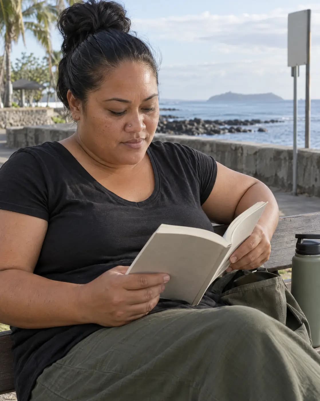 A Pacific Islander woman, 41, broad-shouldered, thick black bun, tiny nose stud reads on a seaside bench