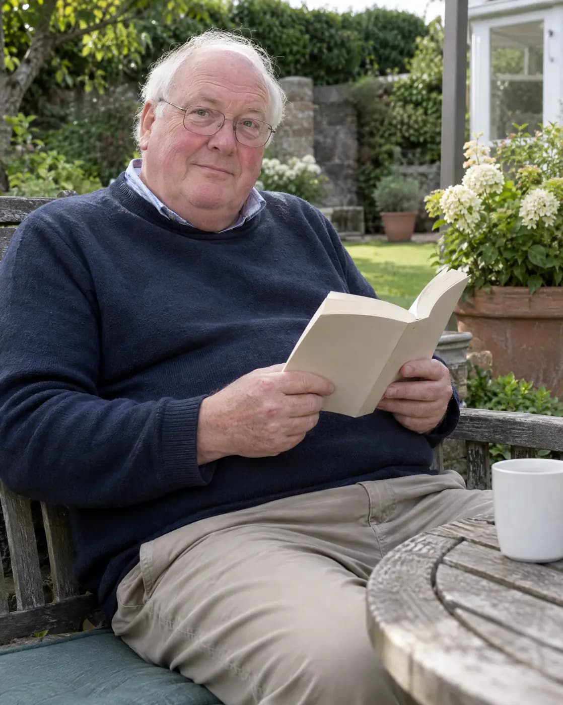 A white man, 63, heavy-set, thin white hair, ruddy cheeks, wire glasses looks up from a paperback on a garden bench