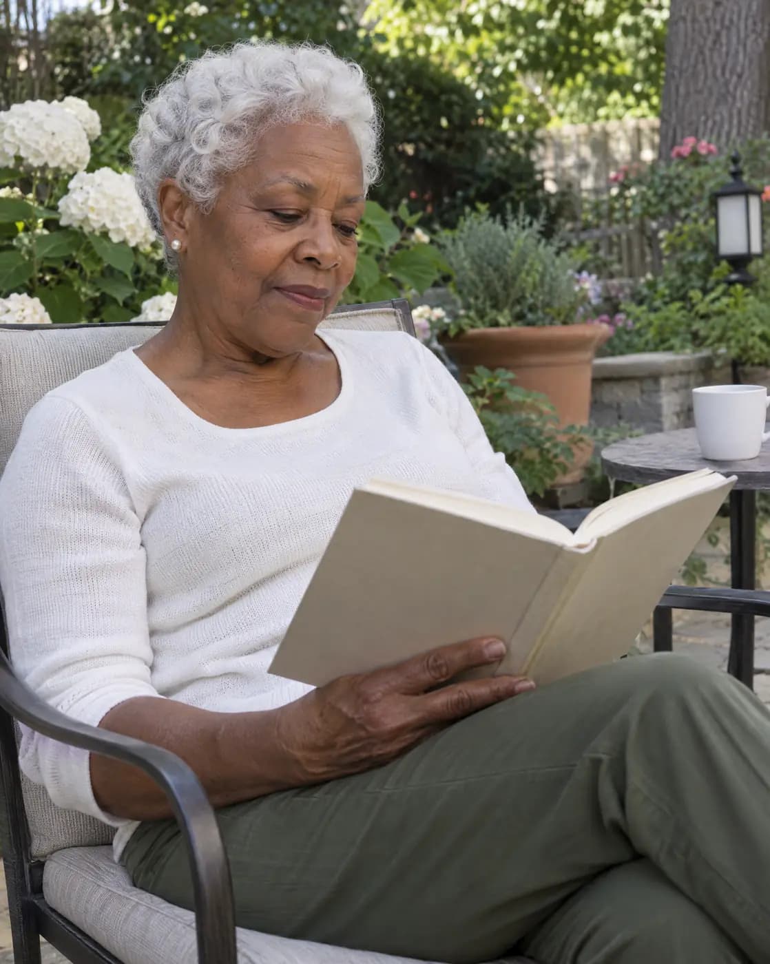 A Black woman, 69, petite, white cropped curls, pearl studs reads in a garden chair
