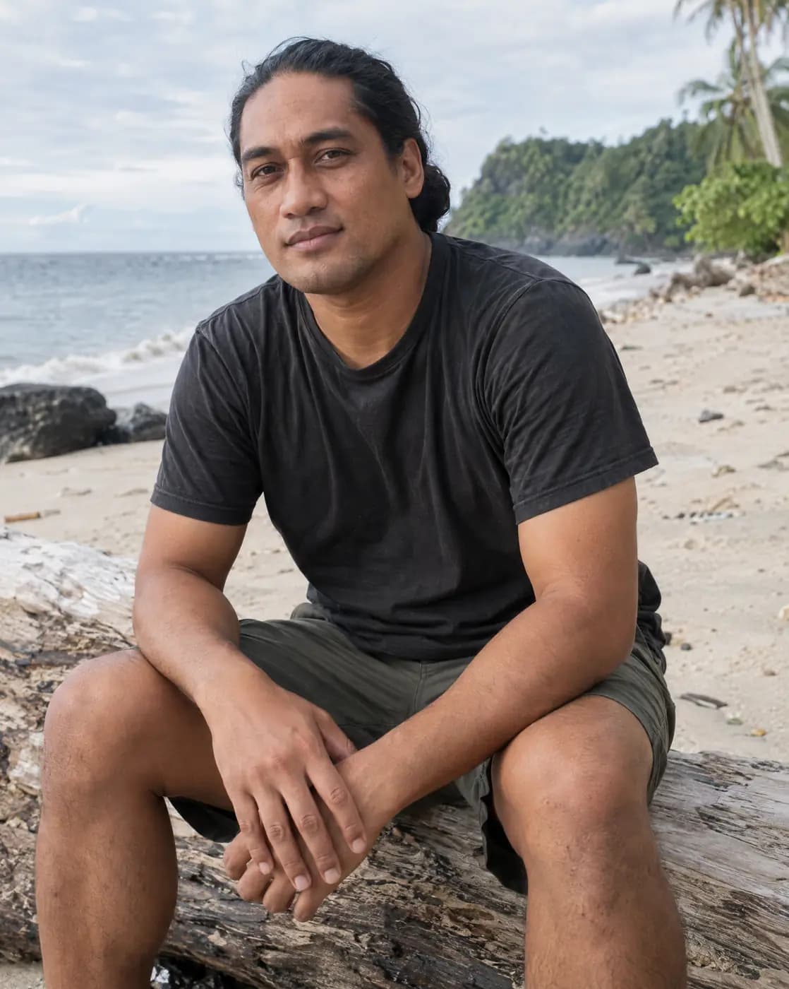 A Samoan man, 38, slim, long black hair tied back, clean shaven sits on a beach log