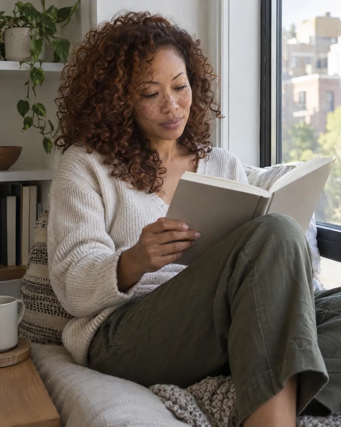 A mixed Black and Asian woman, 42, slim, curly auburn hair, light freckles reads on a windowsill seat