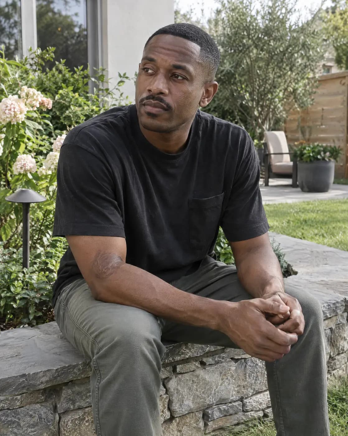 A Black man, 37, lean, close fade, thin moustache, burn mark on forearm sits on a garden wall, thoughtful