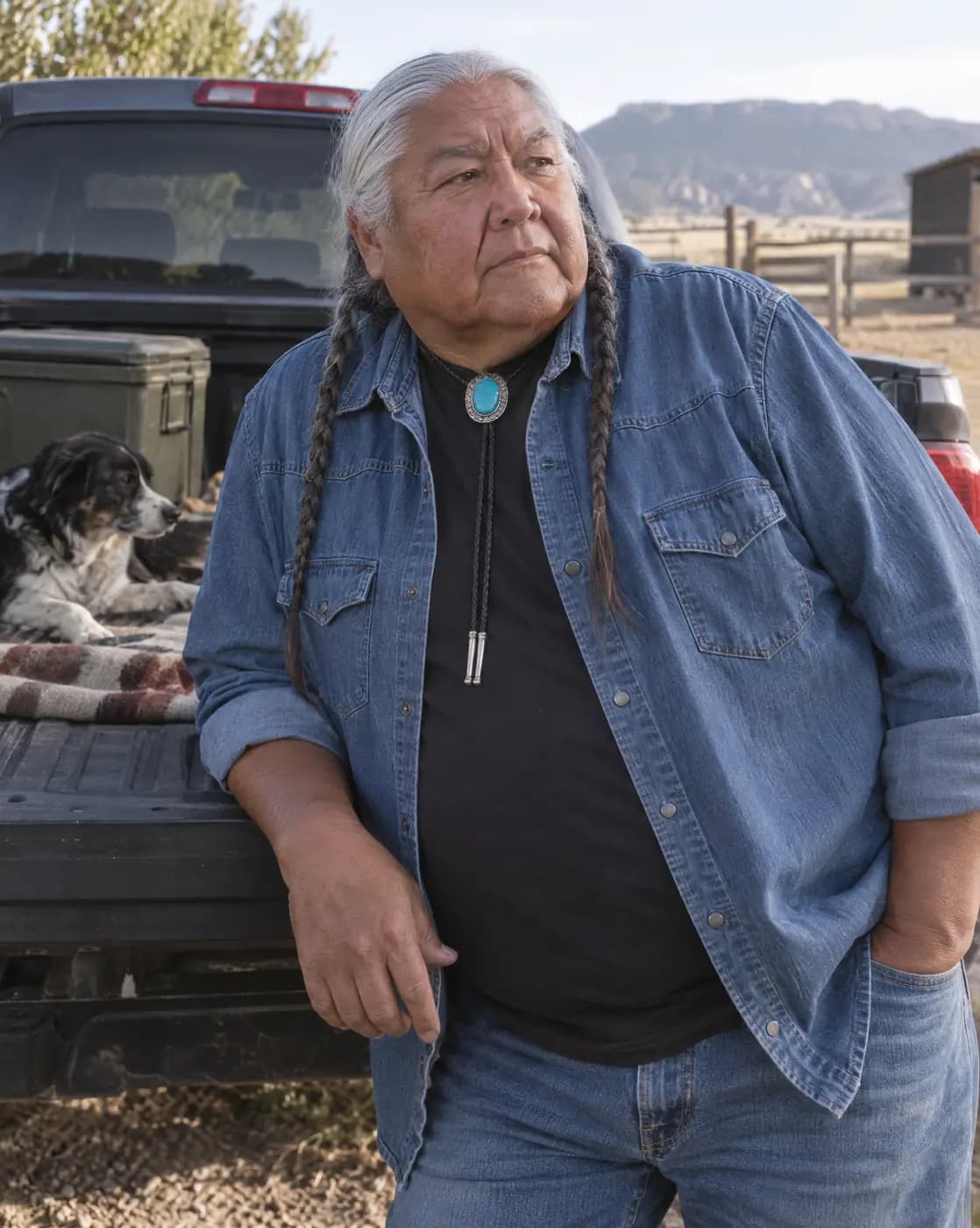 An Indigenous man, 62, heavy, silver braids, turquoise bolo collar rests on a truck tailgate, listening