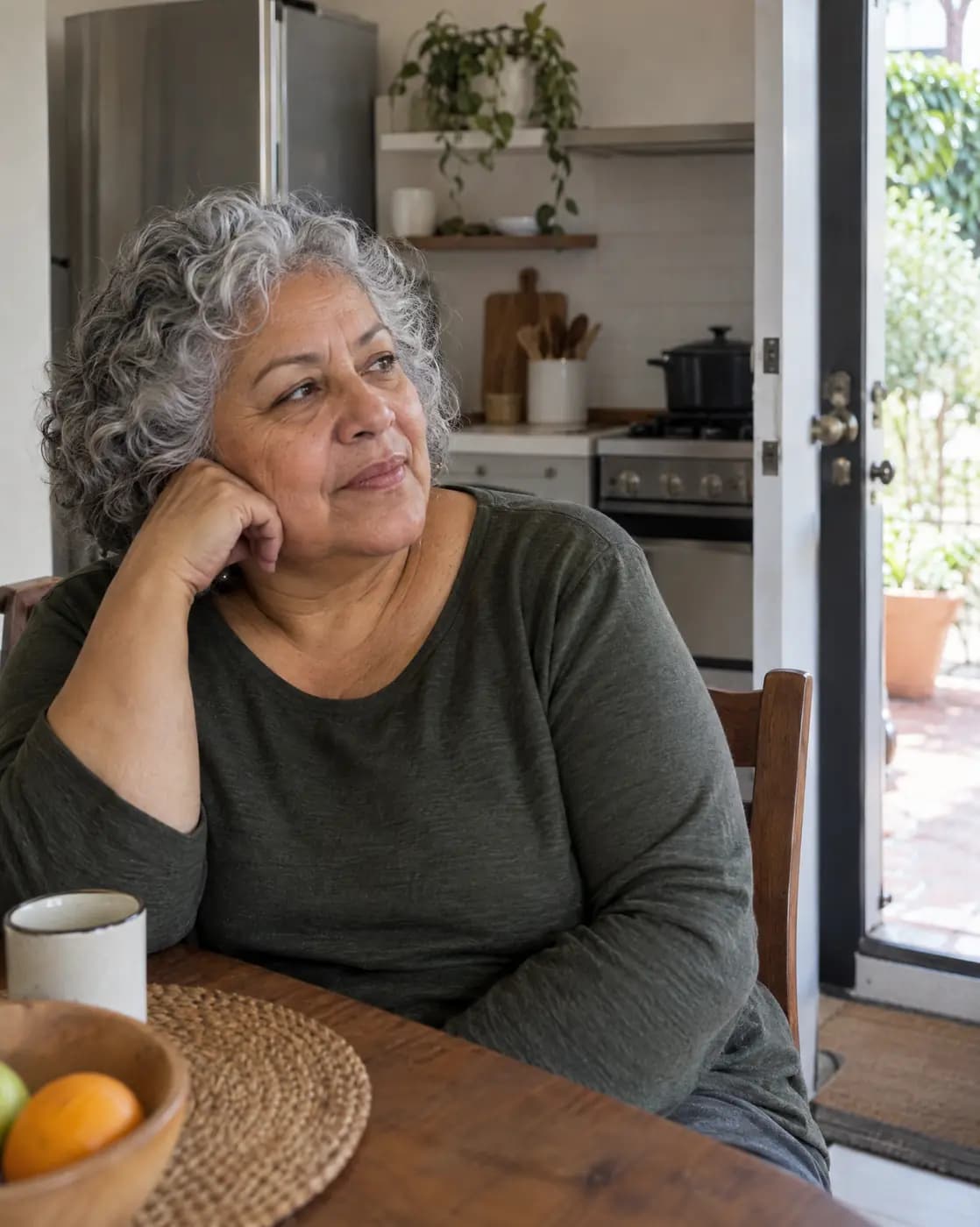 A Latina woman, 65, rounded, silver curly hair, gold earrings sits by an open kitchen door, listening
