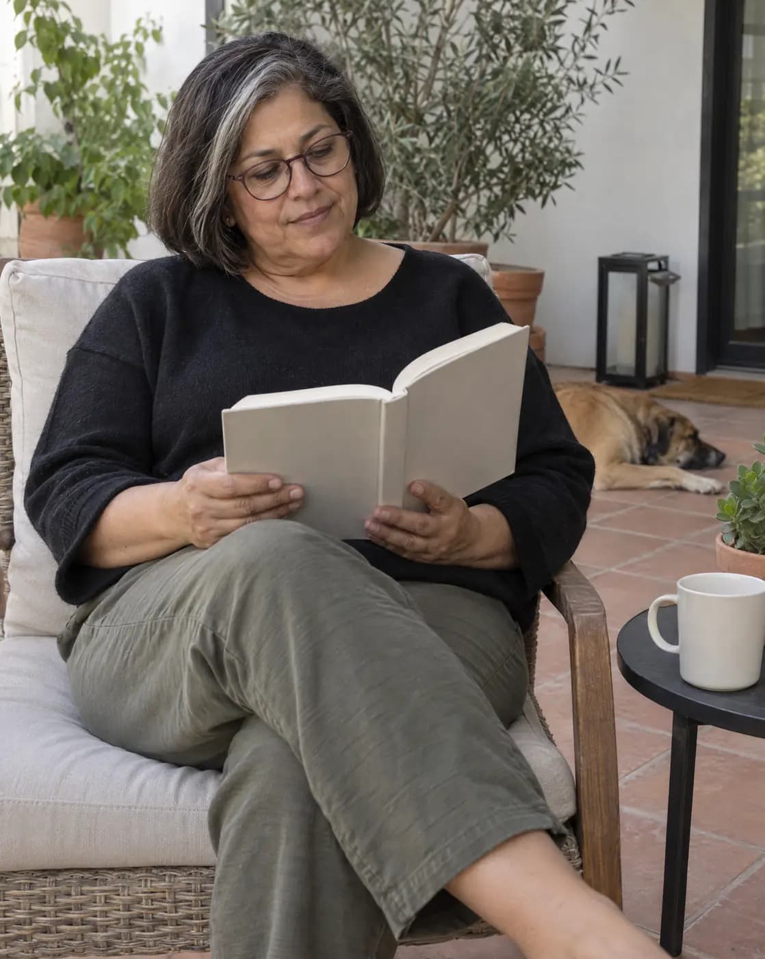A Latina woman, 57, medium build, dark-brown bob with a grey streak, round glasses reads on a tiled patio