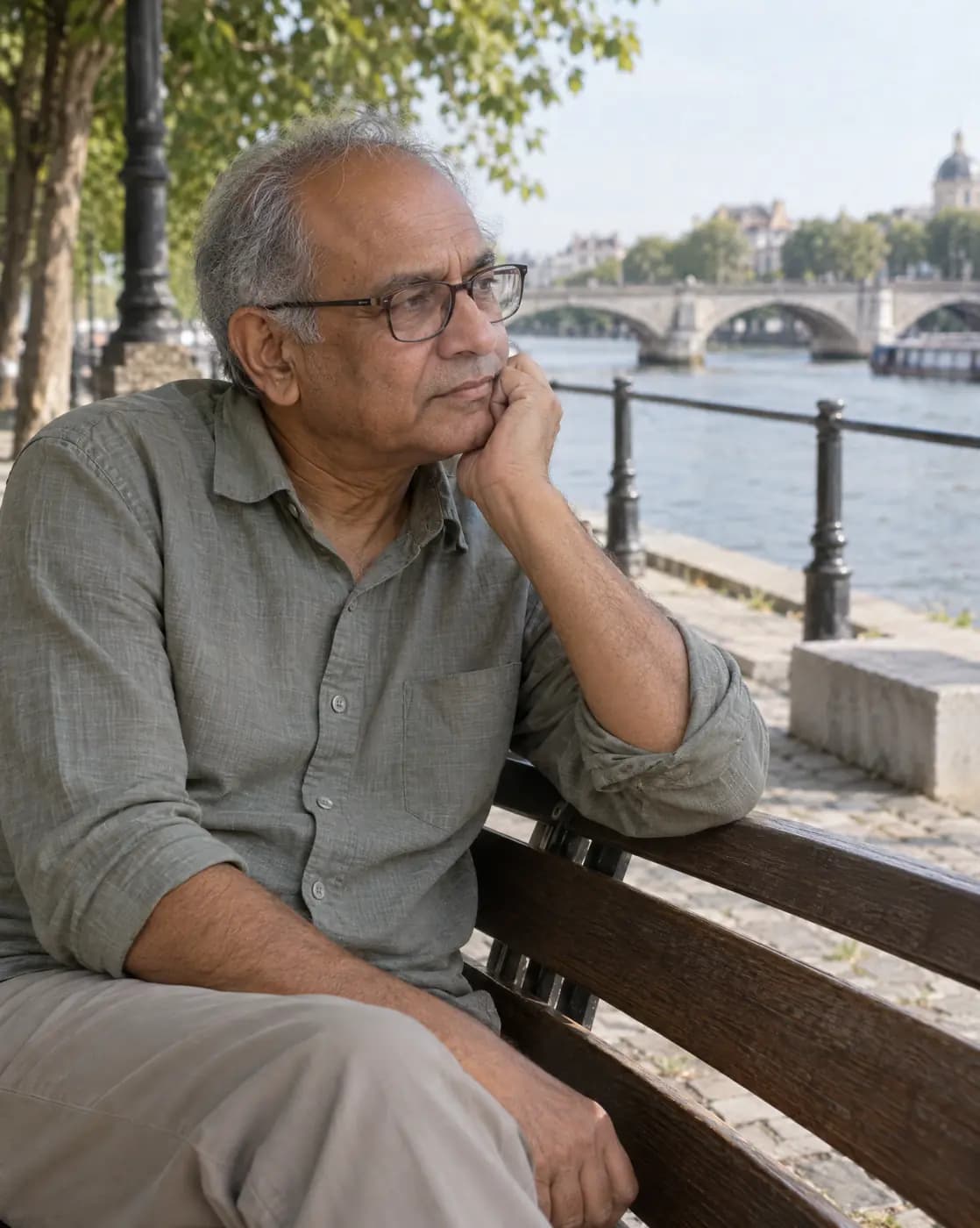 A South Asian man, 60, wiry, thinning grey hair, smooth chin, reading glasses sits at a riverside bench, thoughtful
