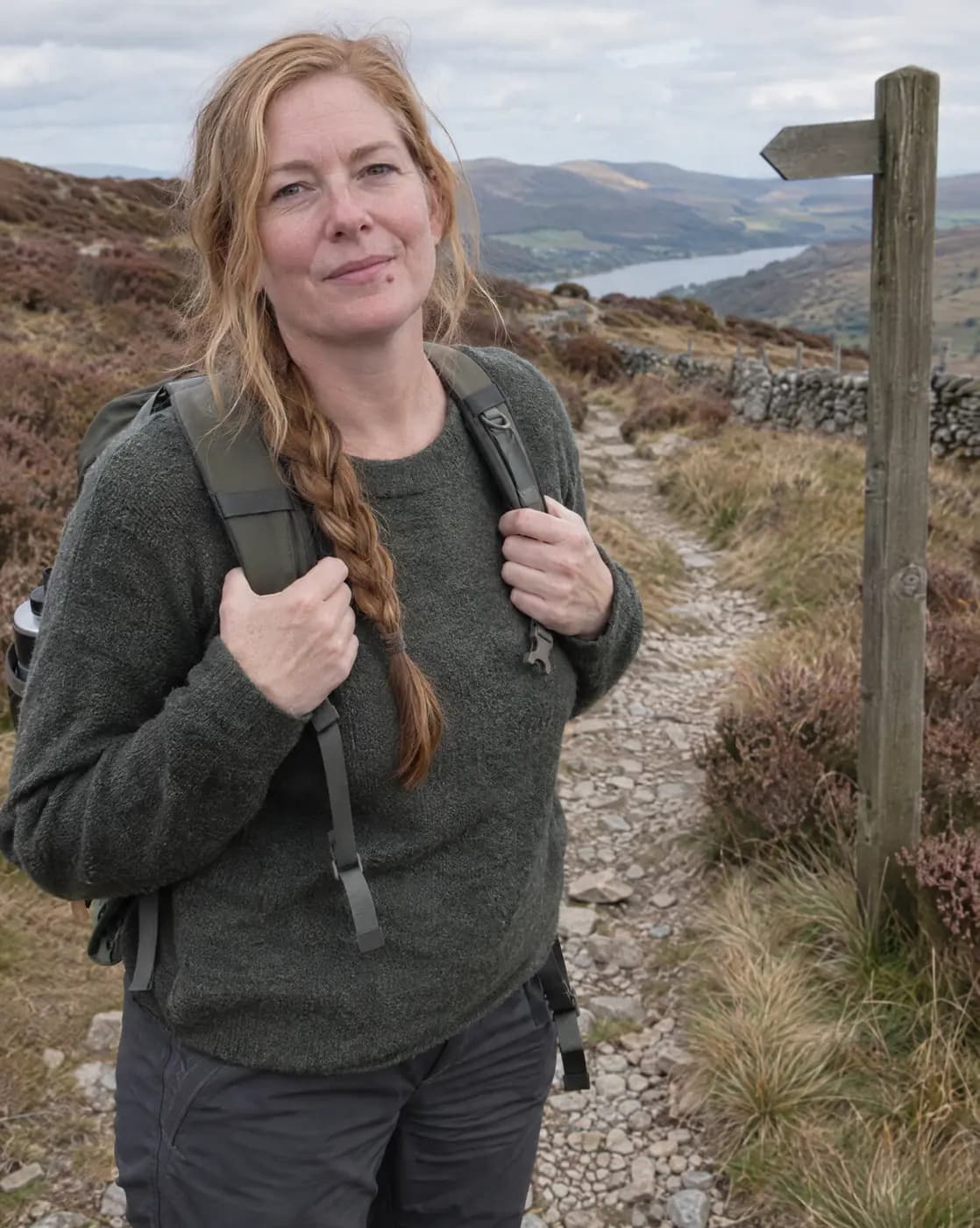 A white woman, 45, tall, strawberry-blonde braid, pale skin, lip freckle pauses on a heather moor path