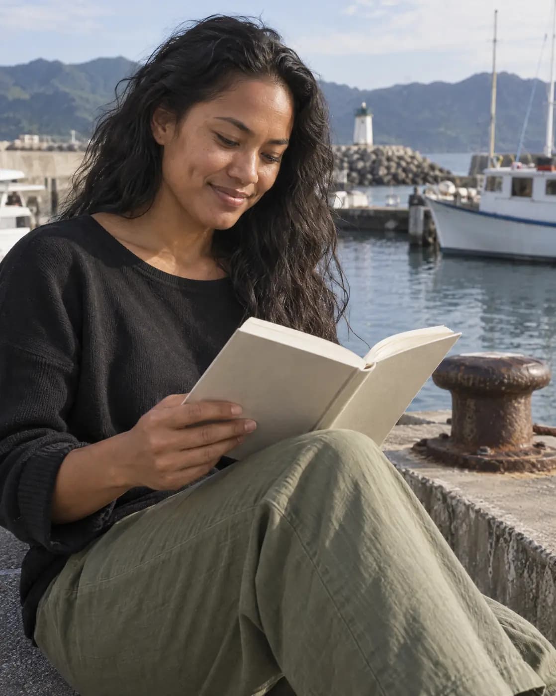 A Pacific Islander woman, 30, slim, wavy black hair, warm brown skin, dimple reads on a harbor wall